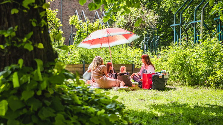2 vrouwen en 2 kinderen picknicken onder een parasol in het gras.