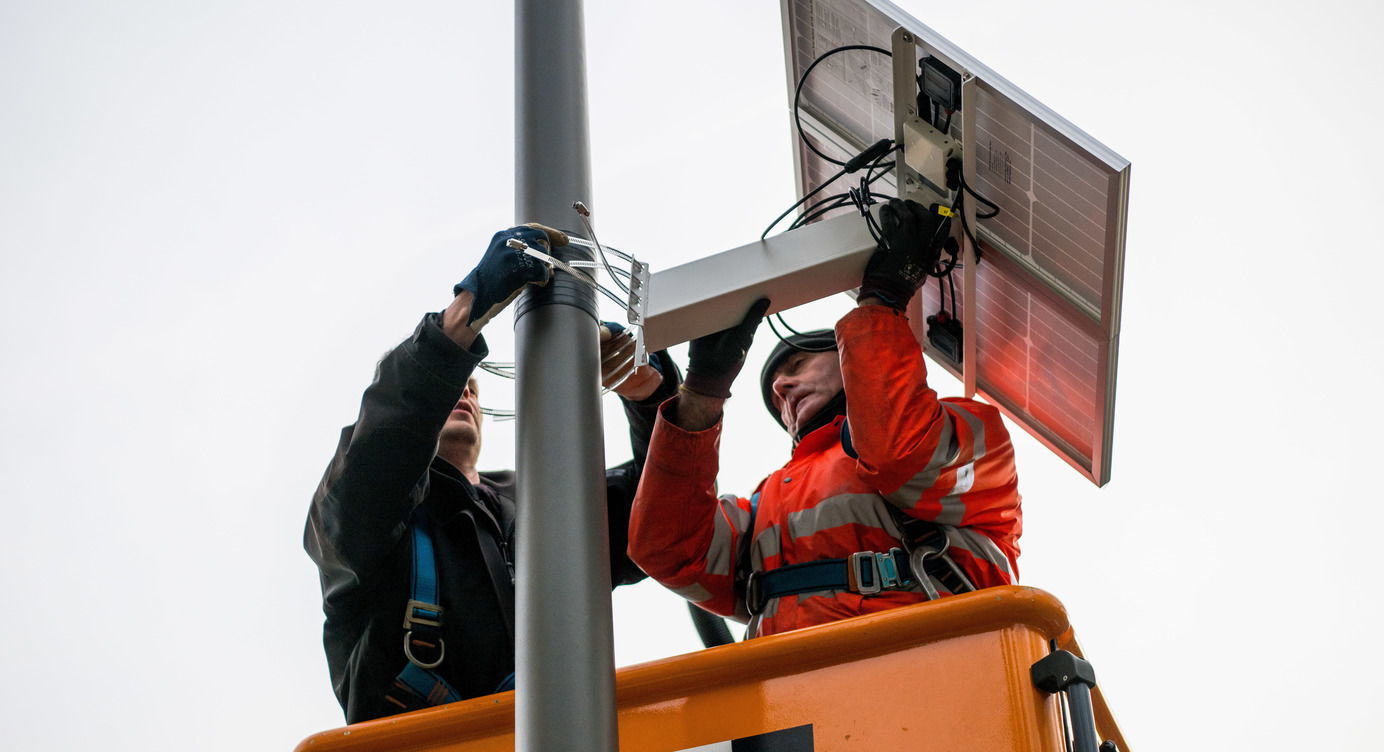 De verkeerstellers worden opgehangen. Twee mannen in een hoogtewerker, installeren een sensor met een zonnepaneel aan een verlichtingspaal.